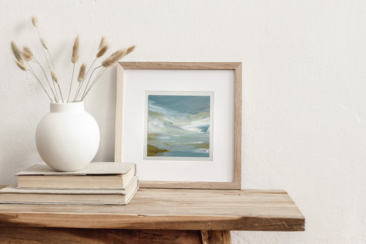Framed artwork on a wooden shelf with a white vase and books